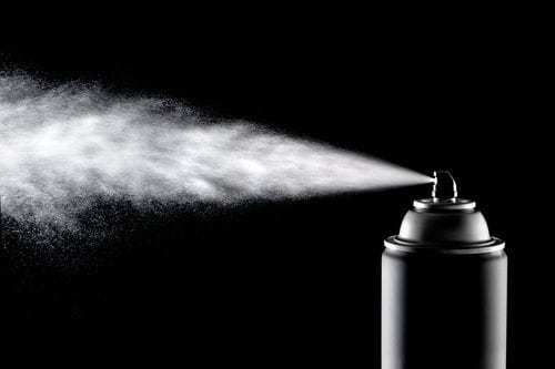 An aerosol can of spray dispensing its content against a backlit black background.